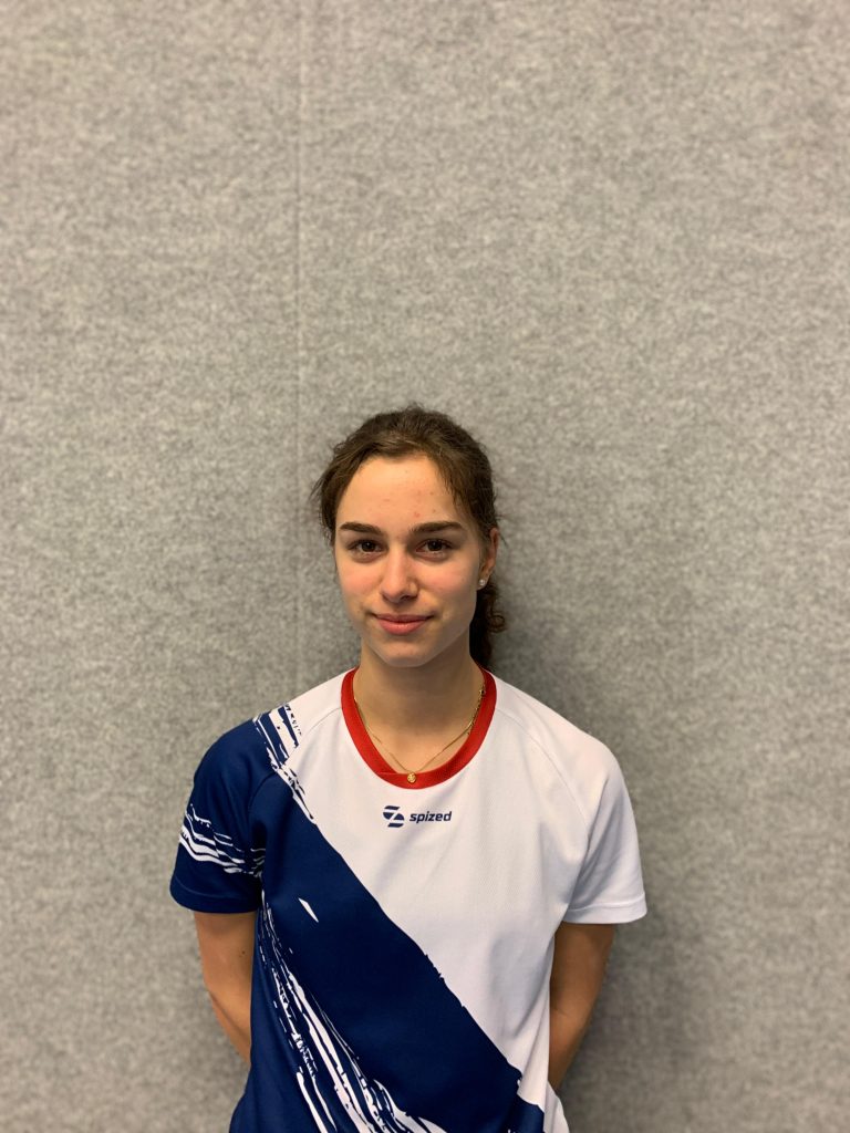 Sportlerin (Amy Rosenthal) mit zusammengebundenem Haar trägt ein weißes Sport-Trikot mit blauem diagonalem Muster und rotem Kragen vor grauer Wand