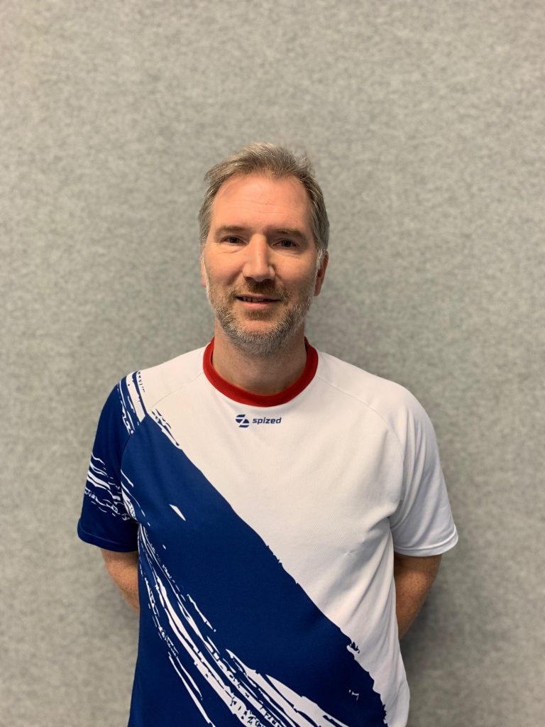 Sportler mit Dreitagebart (Björn Joppien) steht vor grauer Wand und trägt weißes Trikot mit blauem diagonalem Muster und rotem Kragen