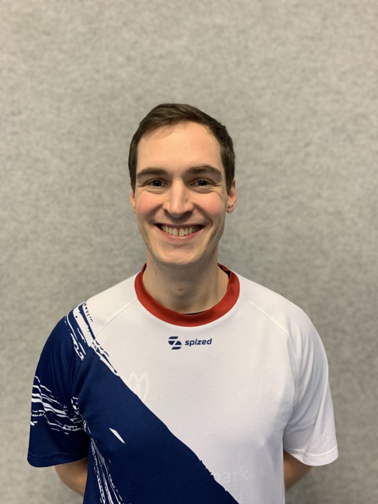 Sportler mit braunem Haar (Christian Müller) steht vor grauer Wand und trägt weißes Trikot mit blauem diagonalem Muster und rotem Kragen