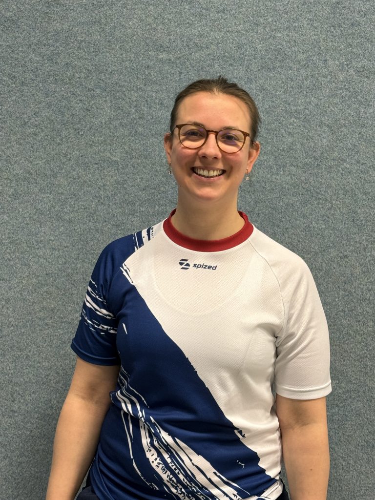 Sportlerin (Elisabeth Duhme) mit zusammengebundenem Haar und Brille trägt ein weißes Sport-Trikot mit blauem diagonalem Muster und rotem Kragen vor grauer Wand