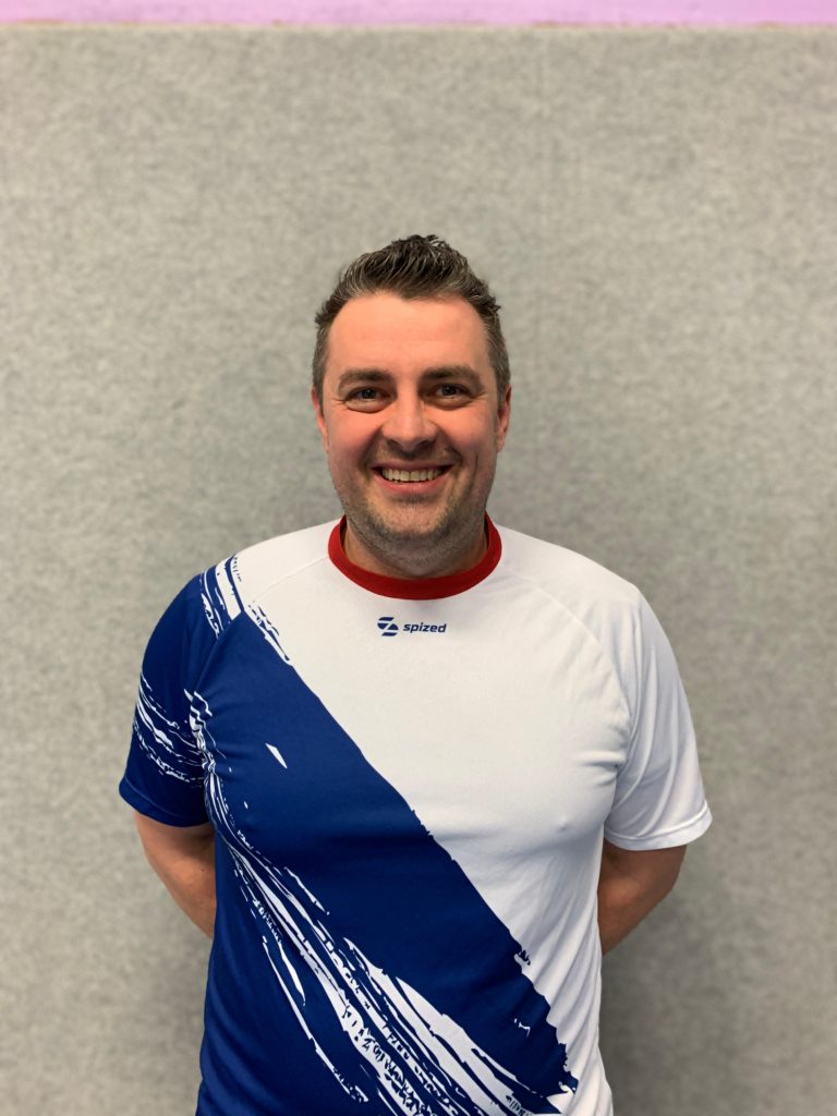 Sportler mit braunem Haar (Jarek Biskup) steht vor grauer Wand und trägt weißes Trikot mit blauem diagonalem Muster und rotem Kragen