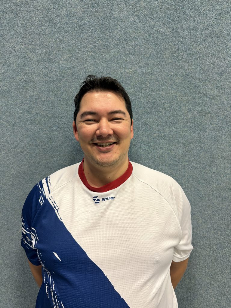 Sportler mit schwarzem Haar (Johannes Bilo) steht vor grauer Wand und trägt weißes Trikot mit blauem diagonalem Muster und rotem Kragen