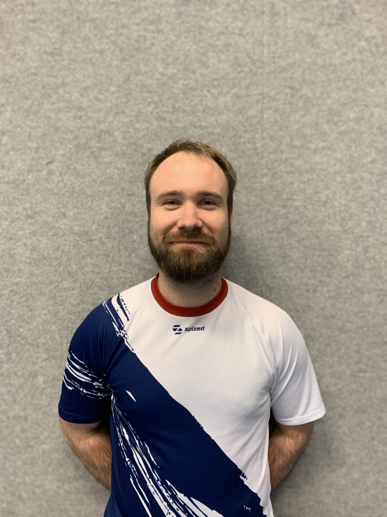 Sportler mit blonden Haaren und Vollbart (Marcel Sieg) steht vor grauer Wand und trägt weißes Trikot mit blauem diagonalem Muster und rotem Kragen
