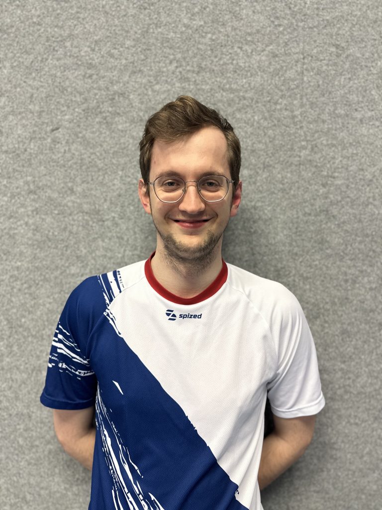 Sportler mit braunem Haar und Brille (Jarek Biskup) steht vor grauer Wand und trägt weißes Trikot mit blauem diagonalem Muster und rotem Kragen