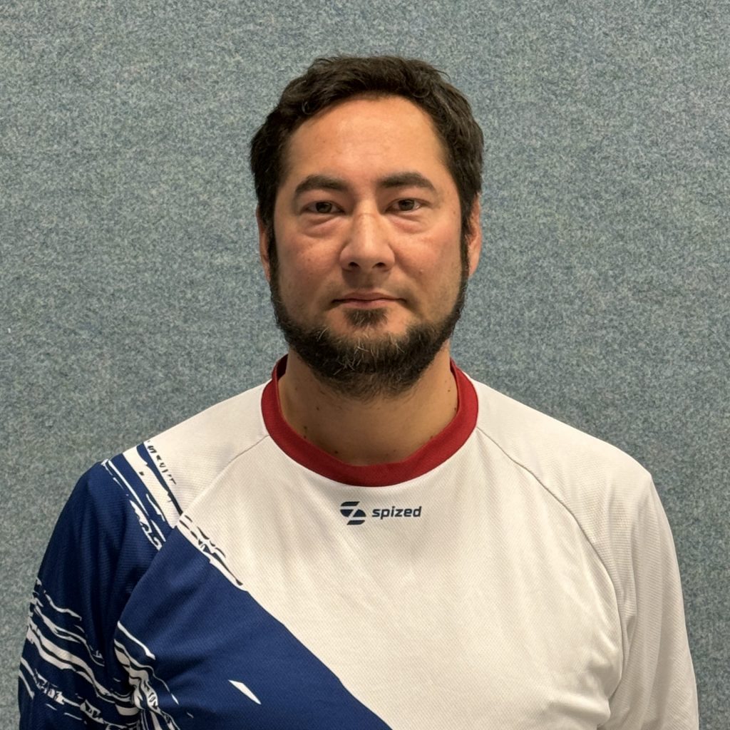 Sportler mit schwarzem Haar und Bart (Matthias Bilo) steht vor grauer Wand und trägt weißes Trikot mit blauem diagonalem Muster und rotem Kragen