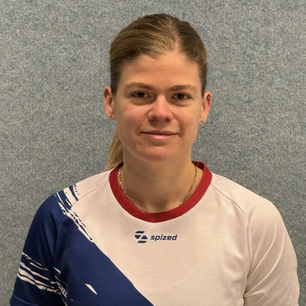 Sportlerin (Nicole Osthoff) mit zusammengebundenem, blondem Haar trägt ein weißes Sport-Trikot mit blauem diagonalem Muster und rotem Kragen vor grauer Wand