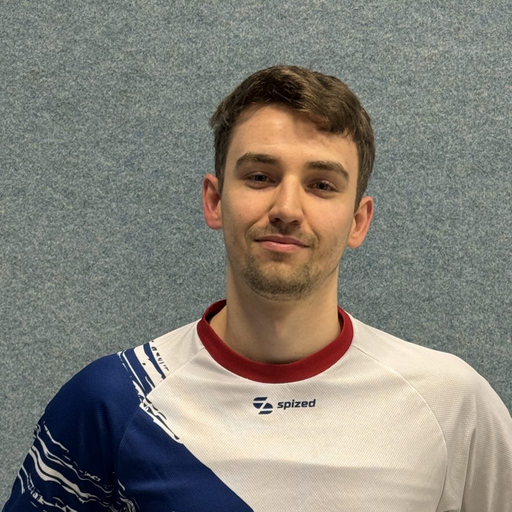 Sportler mit braunem Haar und Dreitagebart (Paul Hofschlaeger) steht vor grauer Wand und trägt weißes Trikot mit blauem diagonalem Muster und rotem Kragen